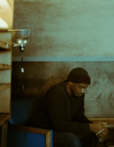 A person wearing a beanie and dark sweater sits in a chair, looking down with hands clasped. Shelves and a lamp are visible beside them.